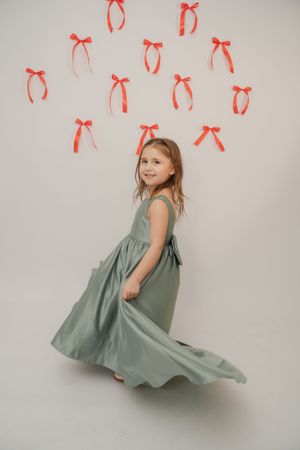 Young girl in sage green dress twirling in studio with red ribbon bows on white wall backdrop.