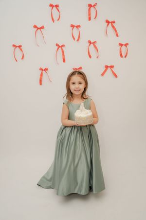 Young girl in green dress holding birthday cake, standing before white wall decorated with red ribbon bows.