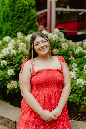 A person in a bright red polka dot sundress stands outdoors near white flowering bushes and greenery.