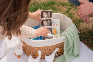 Pregnant couple looking at ultrasound photos in a wicker basket with baby items and rubber ducks on a blanket outdoors.