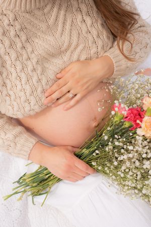 Pregnant woman in beige knit sweater holding colorful flower bouquet, wearing diamond ring, maternity photo.