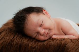 Peaceful newborn baby with dark hair sleeping on a soft brown furry surface against a light blue background.