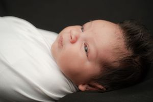 Newborn baby swaddled in white, with dark hair and blue eyes, resting peacefully on a dark background.
