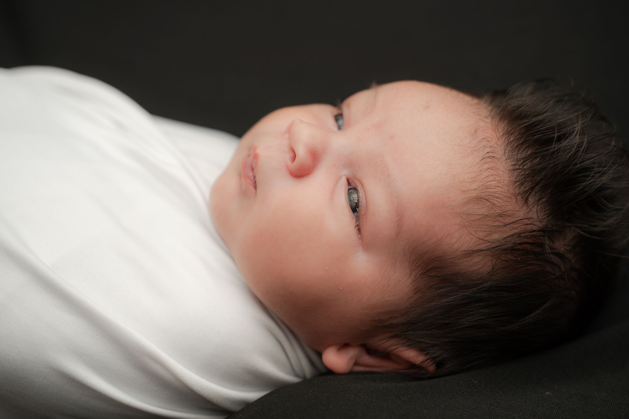 Newborn baby swaddled in white, with dark hair and blue eyes, resting peacefully on a dark background.