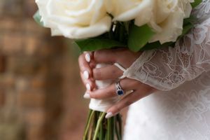 Bride holding white rose bouquet wearing lace wedding dress and sapphire diamond ring on wedding day.