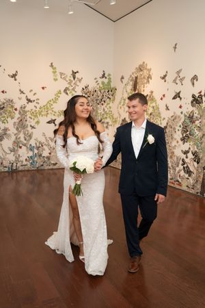 Bride and groom walking hand in hand in an art gallery, bride in white lace gown holding bouquet, groom in navy suit.