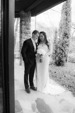 Black and white wedding photo of bride in fitted gown holding bouquet and groom in suit standing together on stone patio.