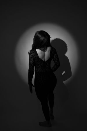 Black and white photo of a woman in a backless lace bodysuit posed under a spotlight with shadow on wall.