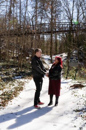 Couple holding hands on a snowy forest trail in winter, wearing black jackets, with a wooden bridge in the background.