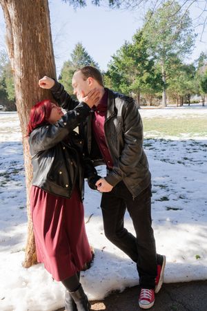 Couple in matching black leather jackets sharing a romantic moment by a tree in a snowy park.