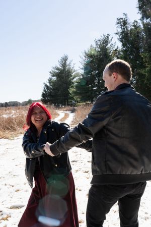 A joyful couple holding hands in a snowy field, wearing black leather jackets, with evergreen trees in the background.
