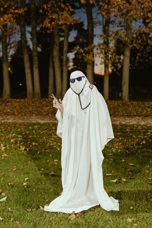 Spooky ghost costume with sunglasses standing on autumn lawn with falling leaves.