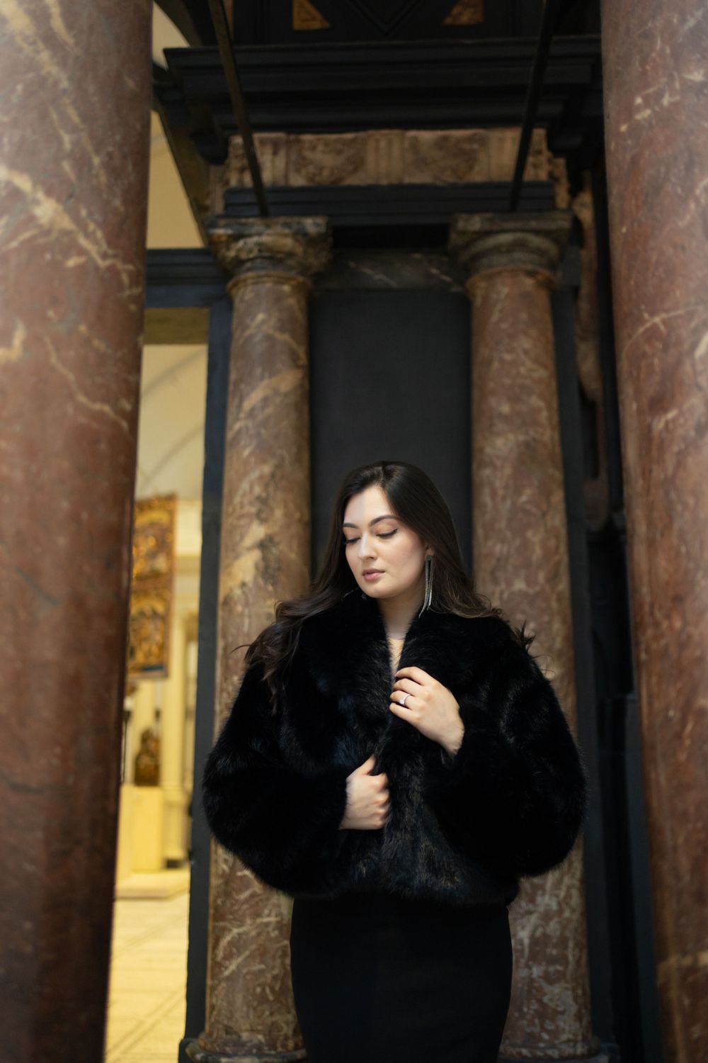 Figure in black coat stands between marble columns in museum hallway.