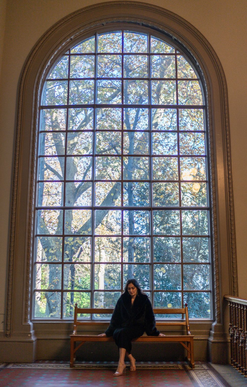 Person in black sits on bench beneath large arched window with natural light streaming in.