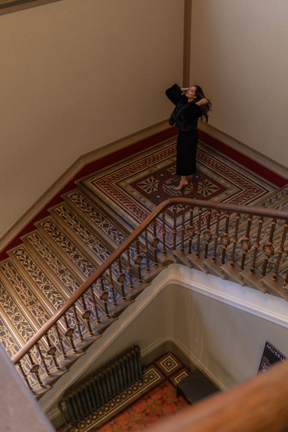Person in black dress poses on ornate vintage staircase with decorative railings.
