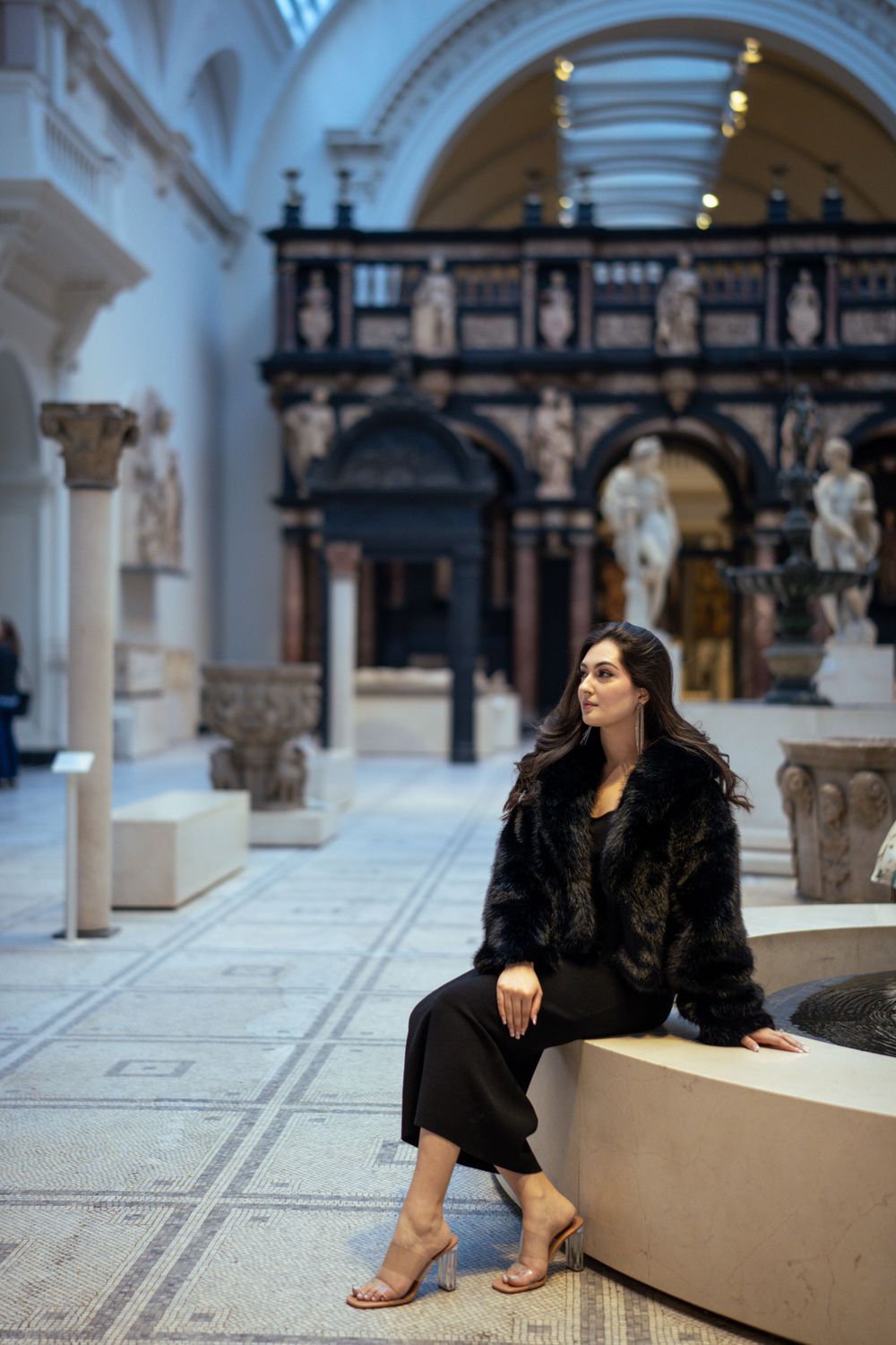 Fashion model in black fur coat poses elegantly in ornate museum gallery with classical architecture and statues.