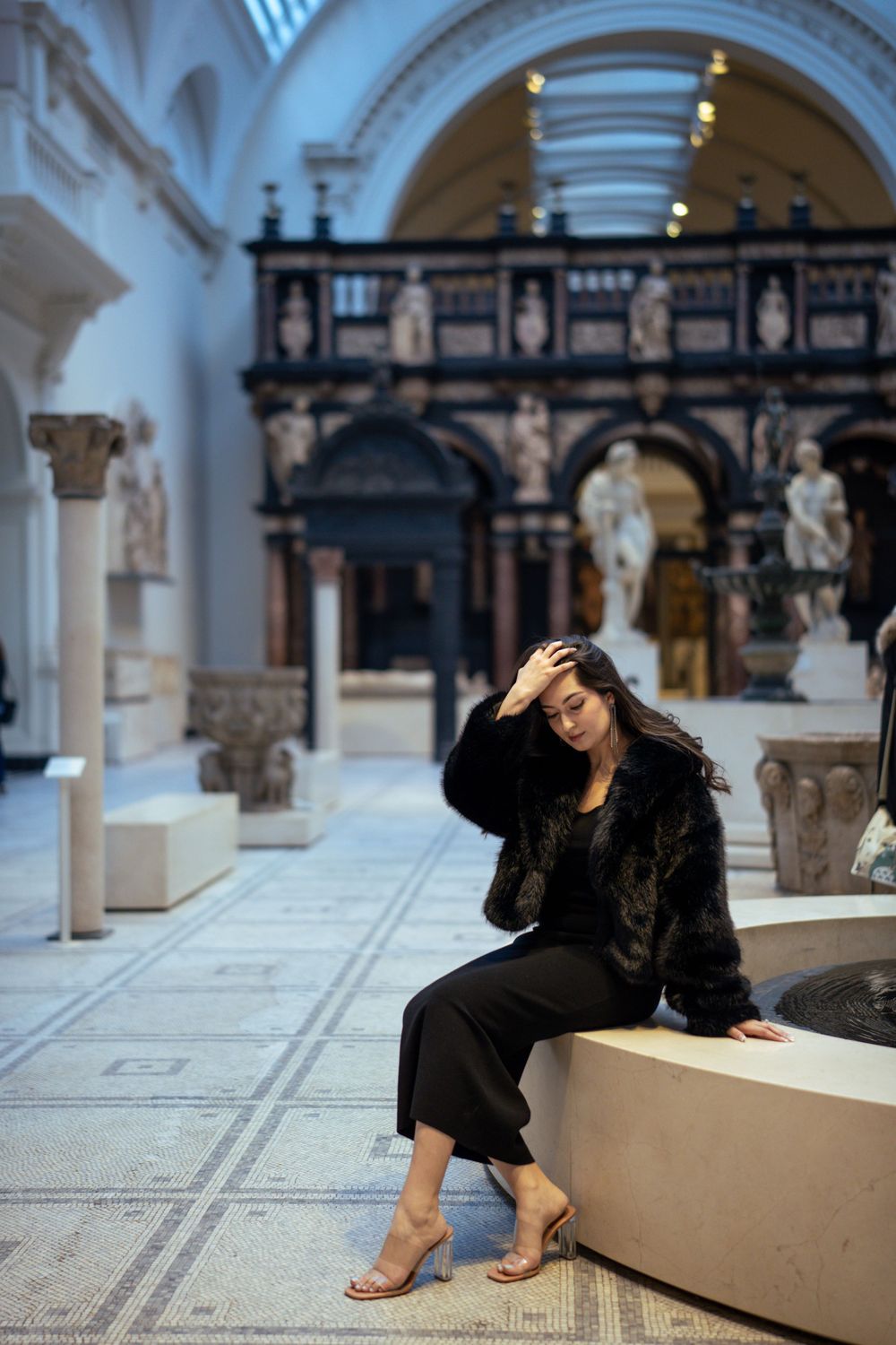 Person in black outfit sits on museum bench surrounded by classical architecture and statues.