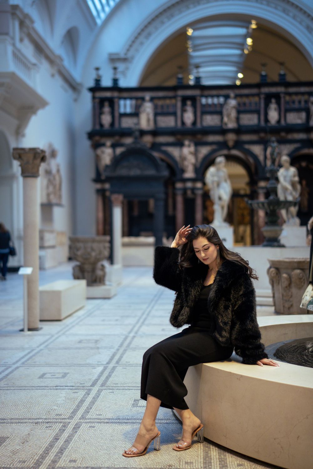 Model poses on museum bench surrounded by classical architecture and marble sculptures.