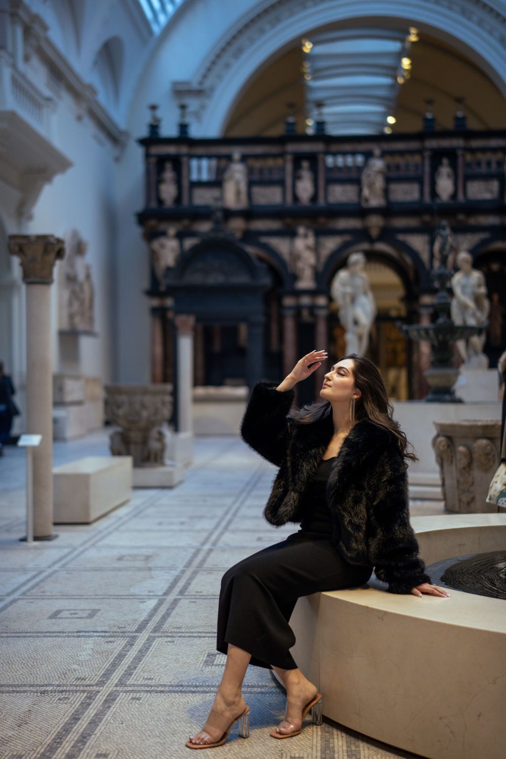 Stylish portrait in museum gallery featuring black fur coat and heels against classical backdrop.