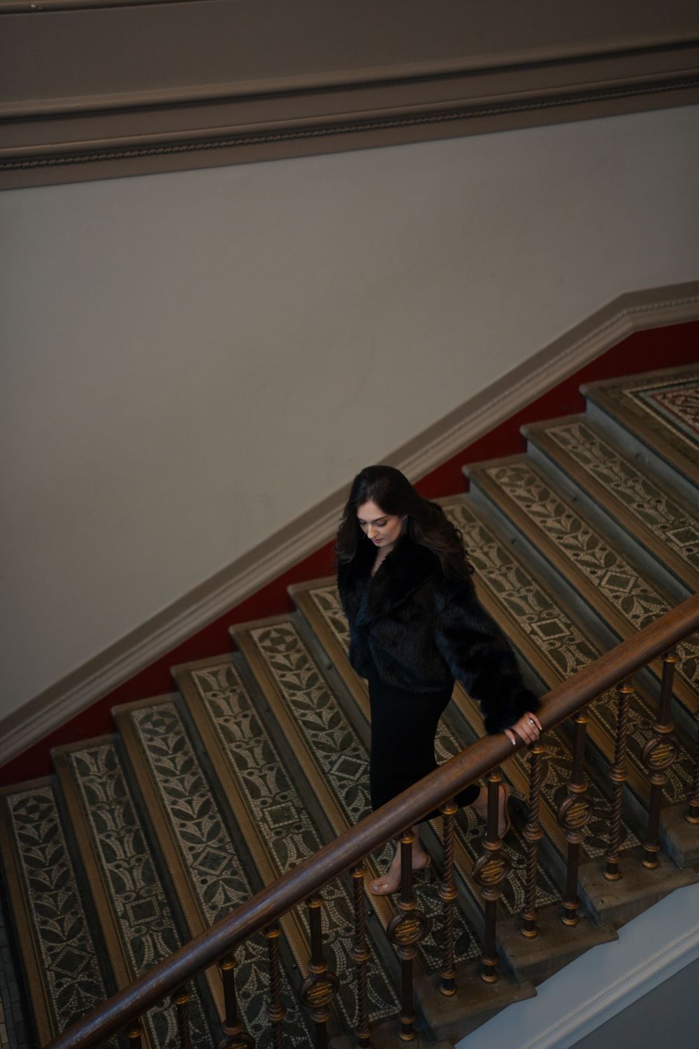Elegant figure descends ornate vintage staircase wearing black fur coat in moody lighting.