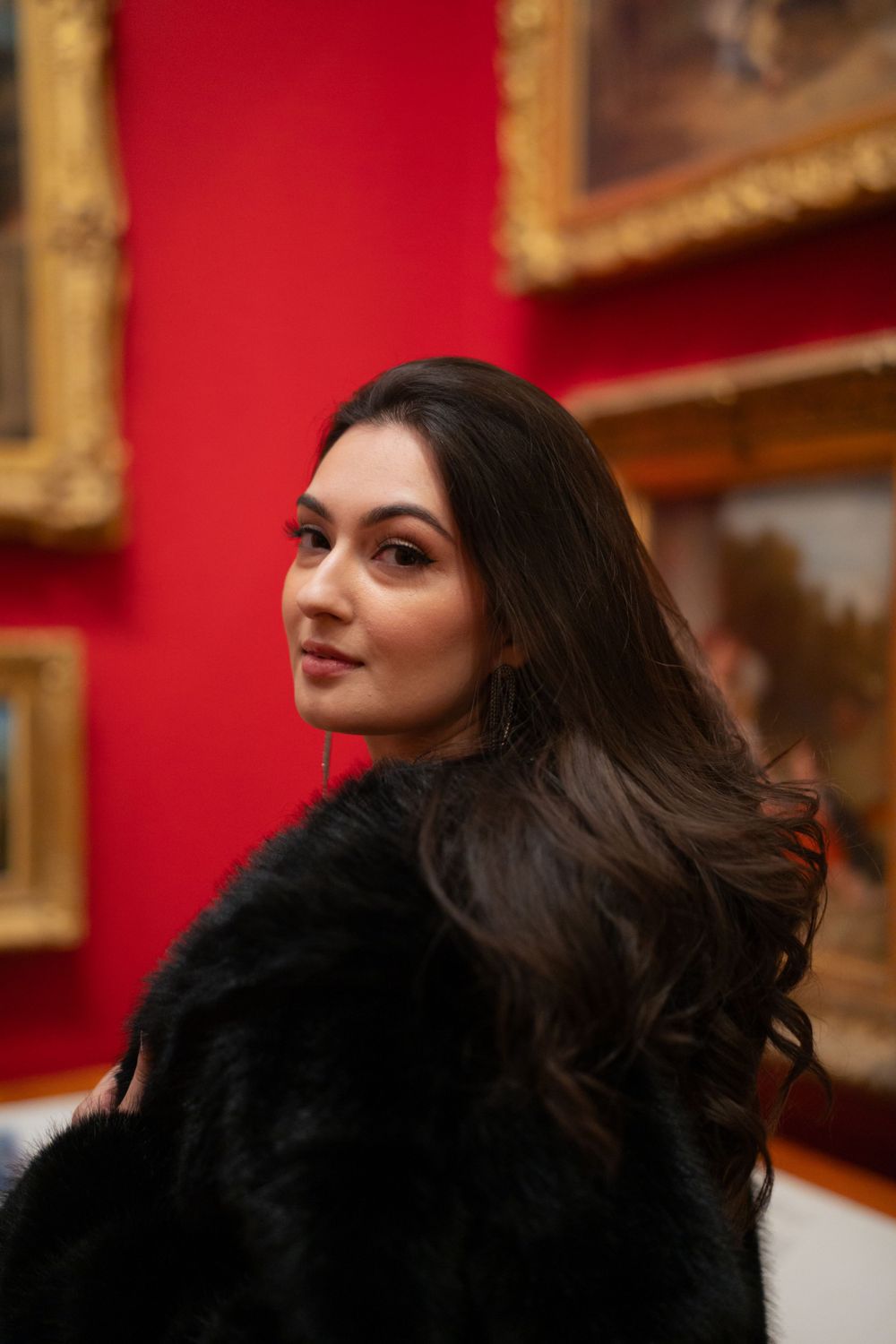 Model wearing black fur coat poses in art gallery with red walls.