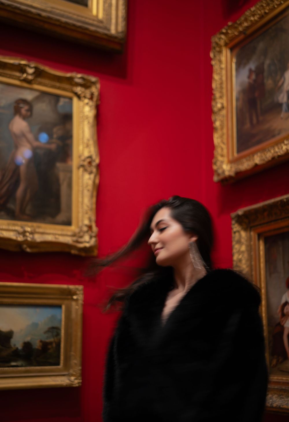 Figure in black coat poses against red gallery wall with ornate gold-framed paintings.