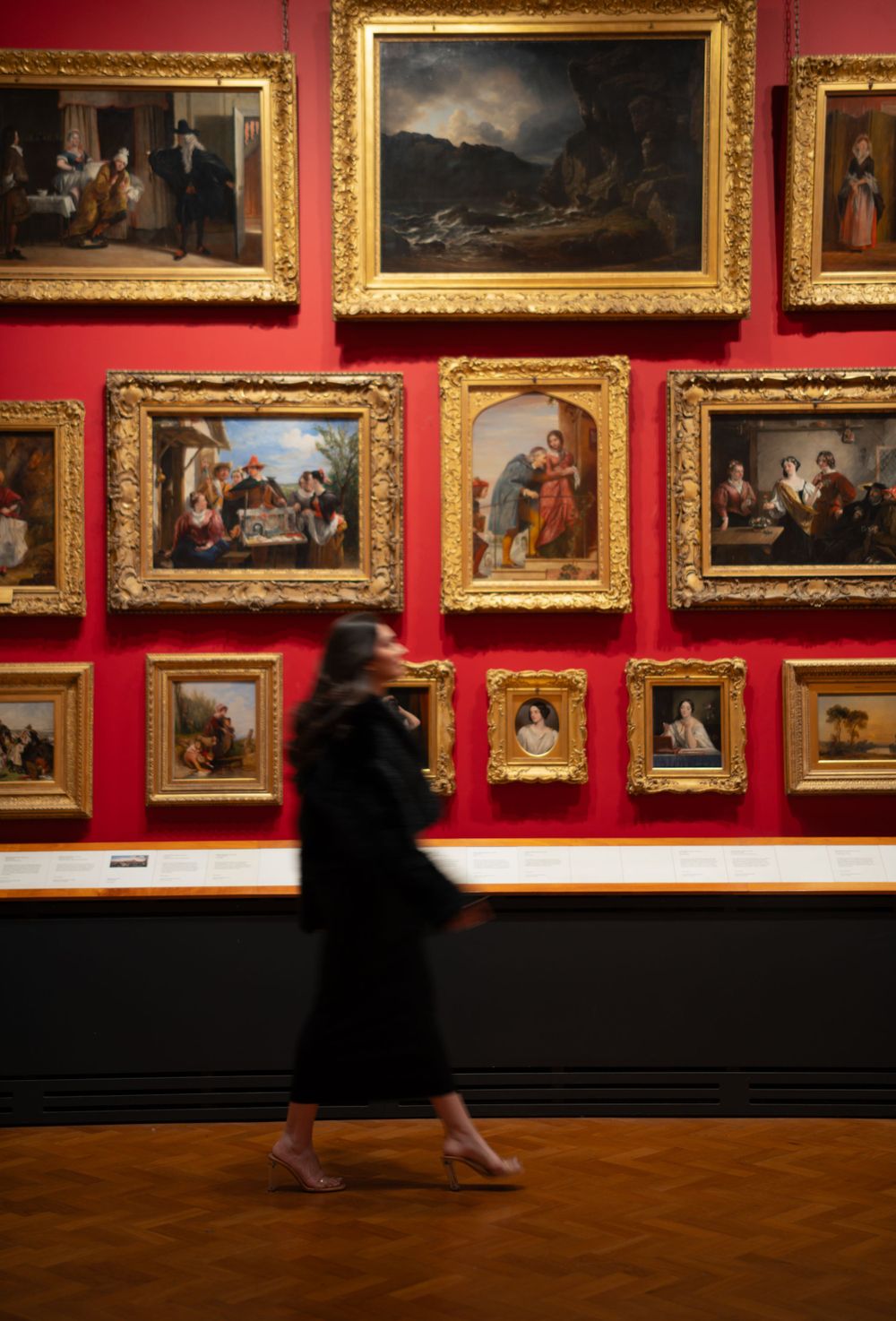 Art gallery visitor walks past classical paintings displayed on red walls with ornate gold frames.