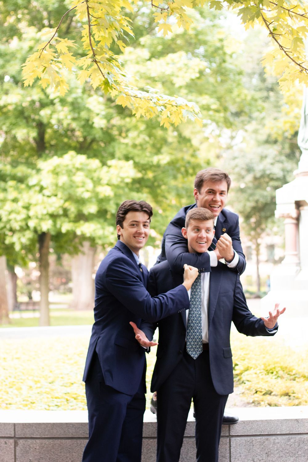 Two people in navy suits playfully posing together outdoors with autumn trees in the background.