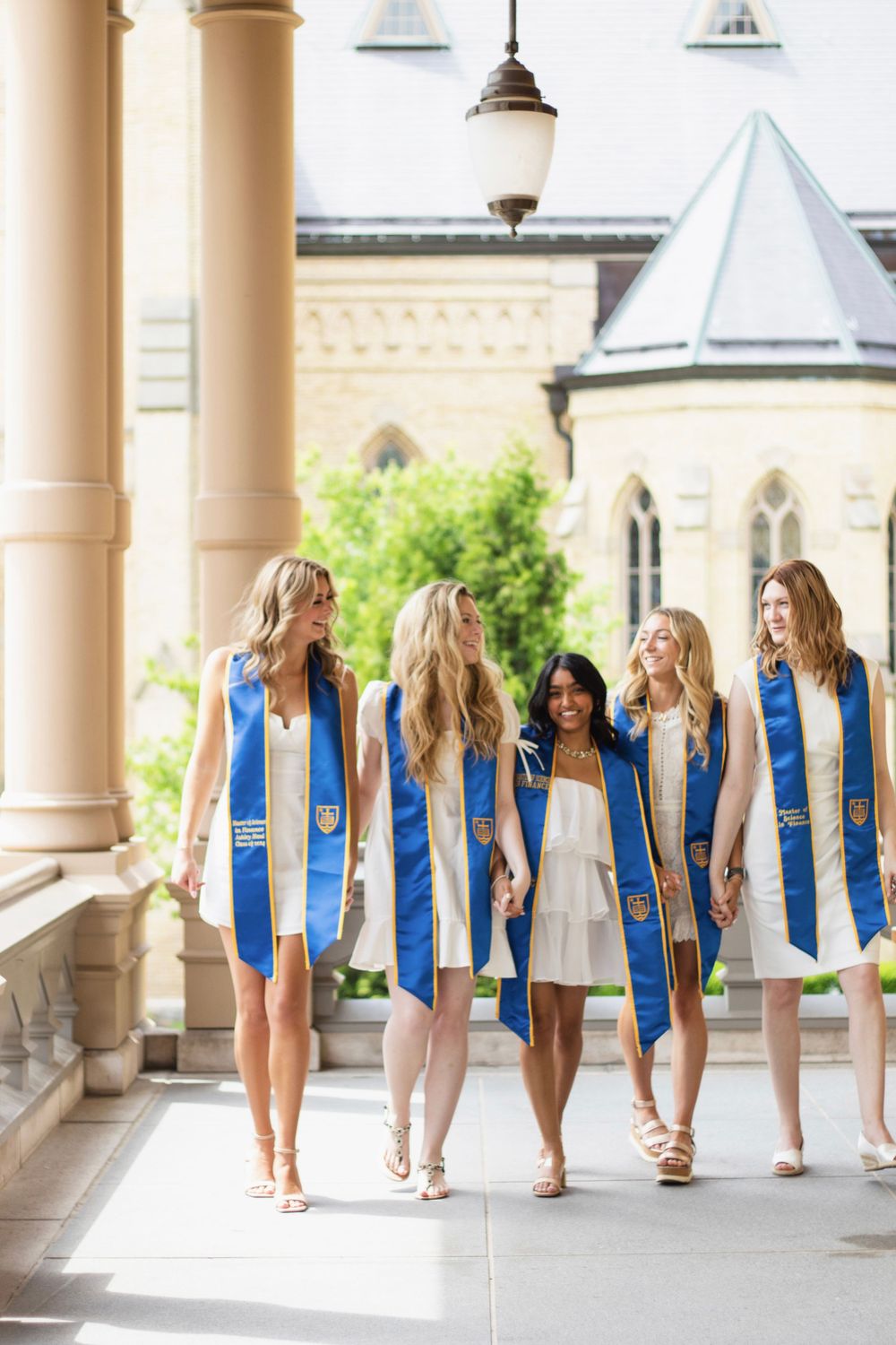 Graduates in blue and white stoles walking together under elegant stone archways.