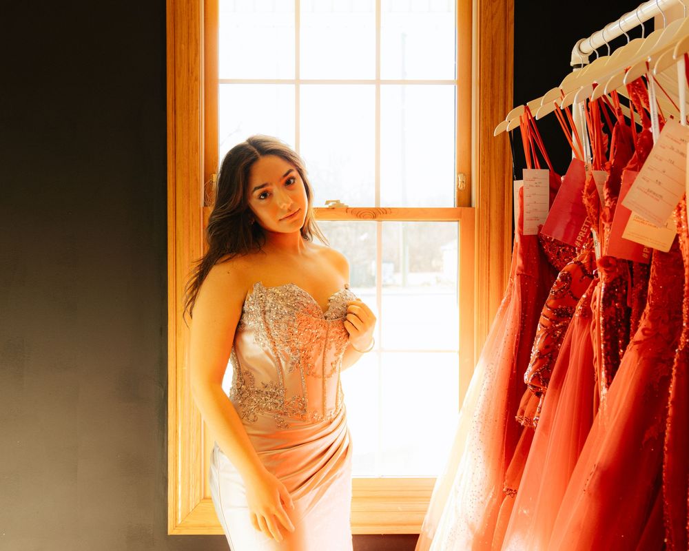 A sparkly champagne-colored formal dress displayed near a window with red dresses hanging in background.