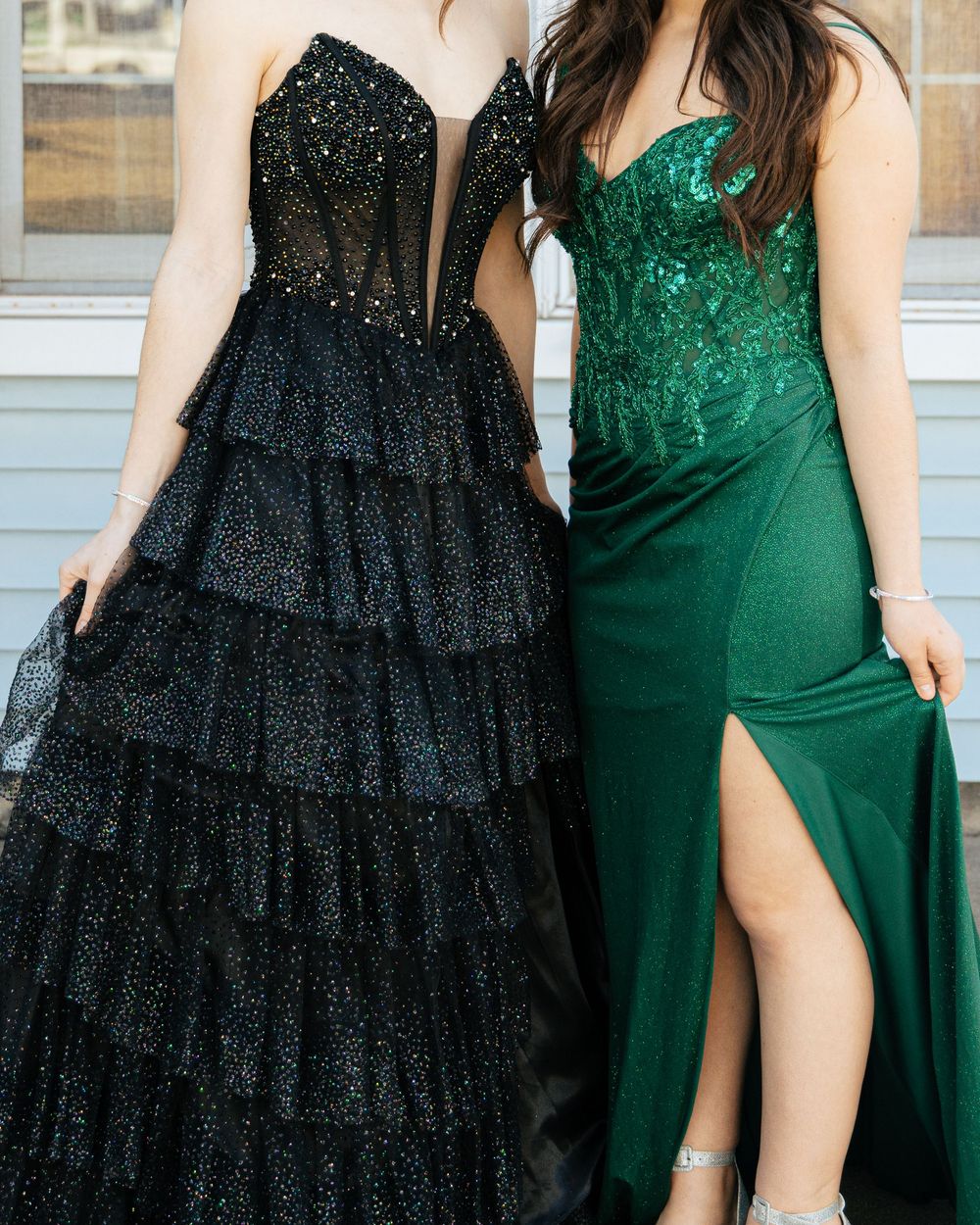 Two elegant evening gowns side by side, one black tiered sparkly dress and one emerald green dress with a thigh slit.