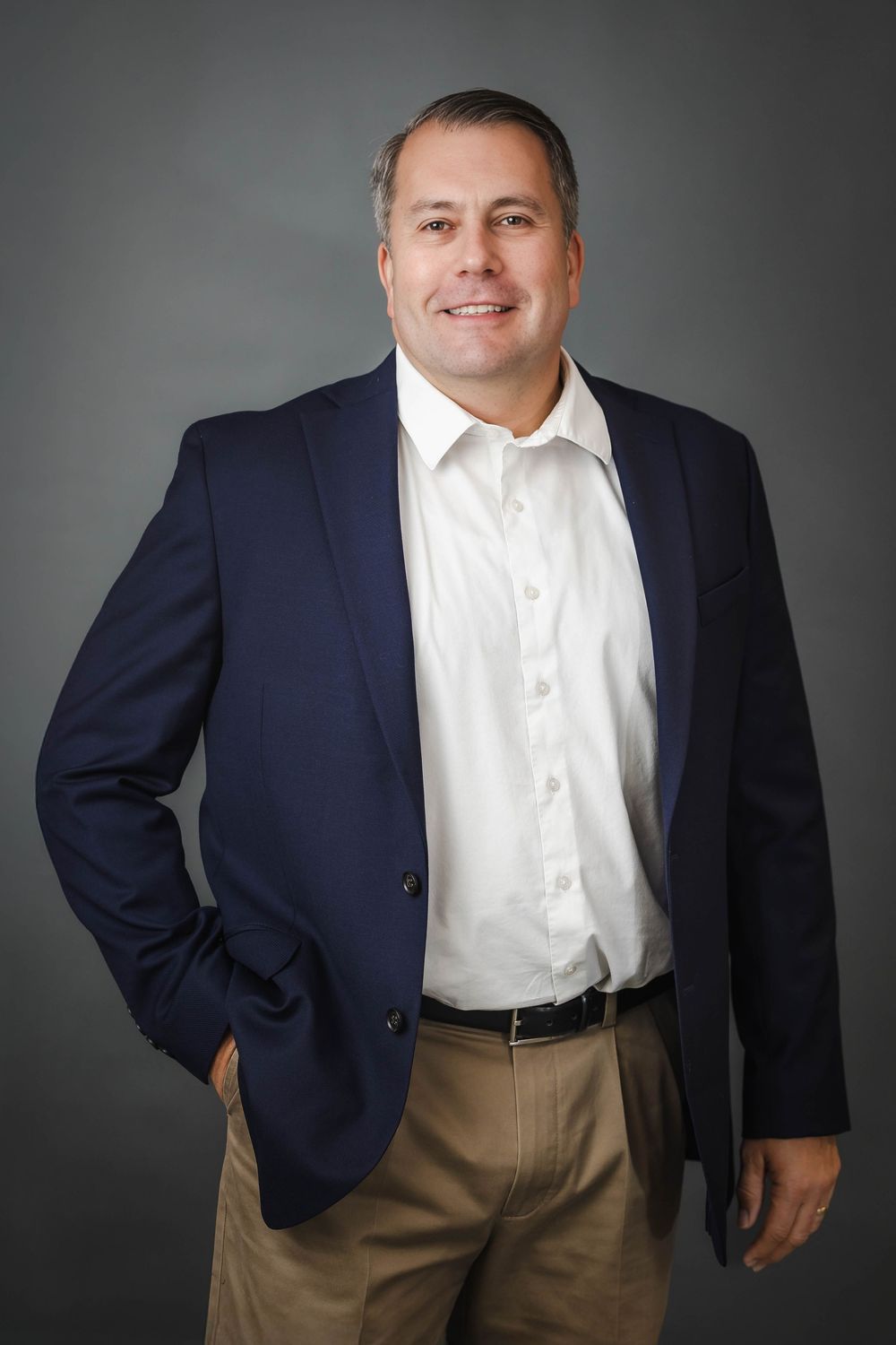 Smiling businessman in navy blazer and white shirt posing casually against a gray studio background.