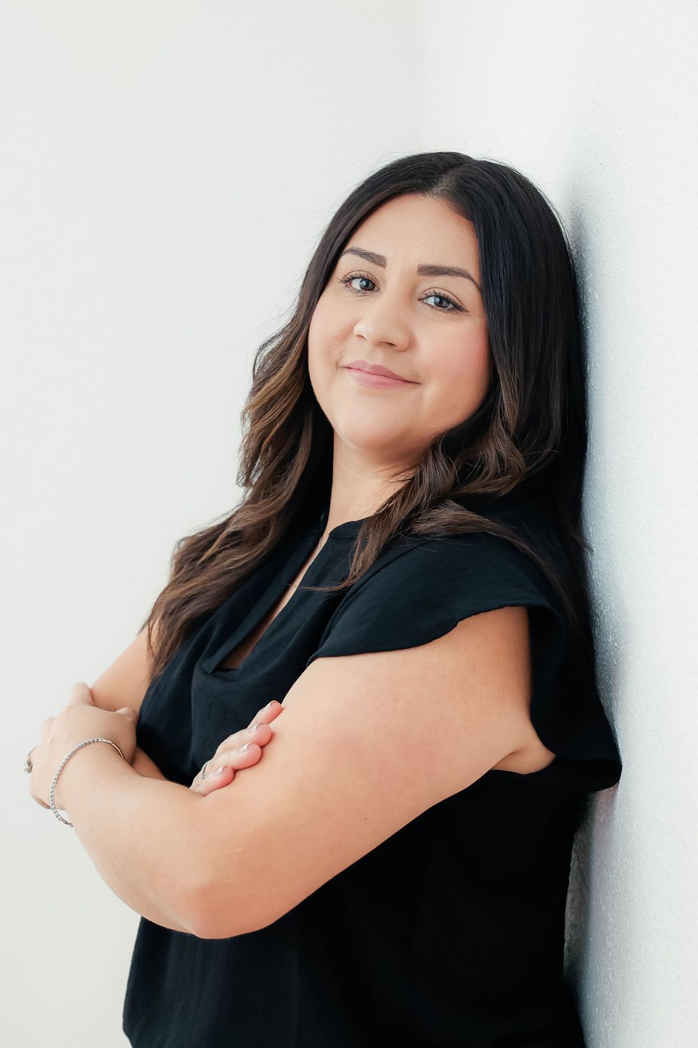 Brunette woman in black top leaning against white wall with arms crossed, smiling.
