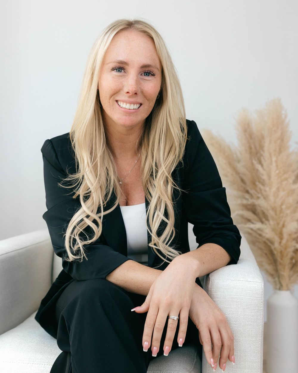 Smiling blonde woman in black blazer seated on white chair with pampas grass decor.