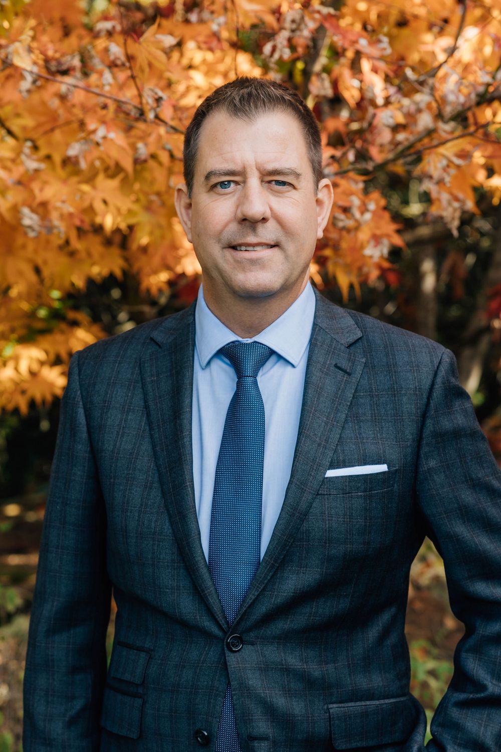 Professional man in dark suit and blue tie stands before vibrant autumn foliage.