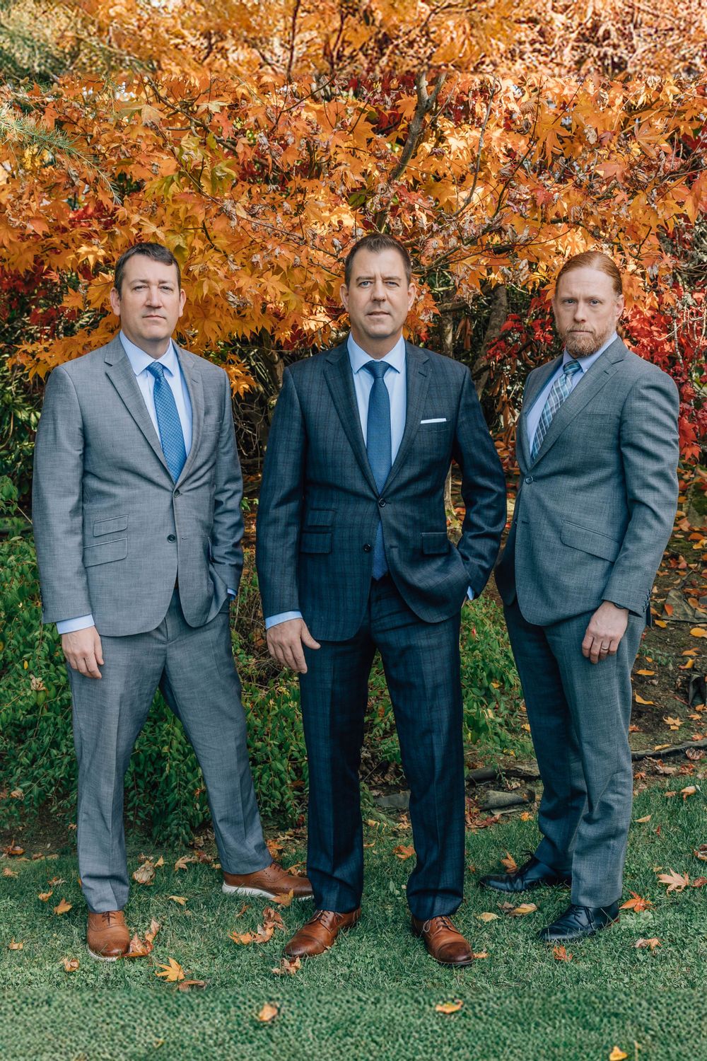 Three businessmen in suits pose seriously before colorful fall trees outdoors.