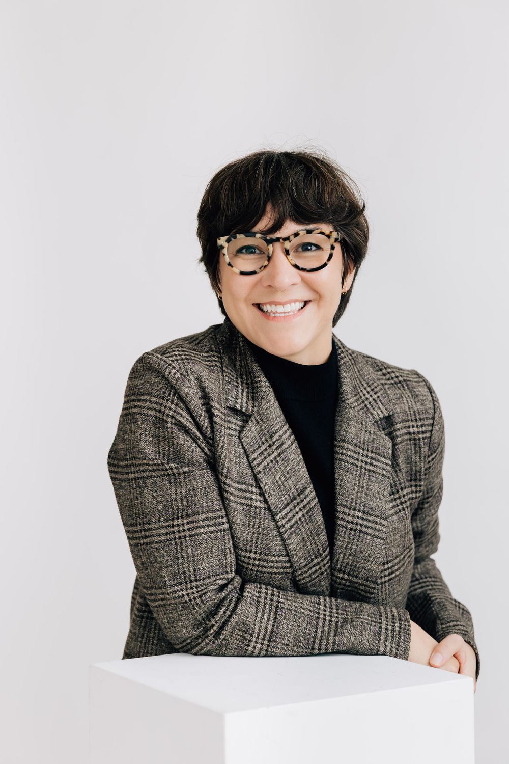 Professional headshot of smiling woman in plaid blazer and glasses leaning on white pedestal.