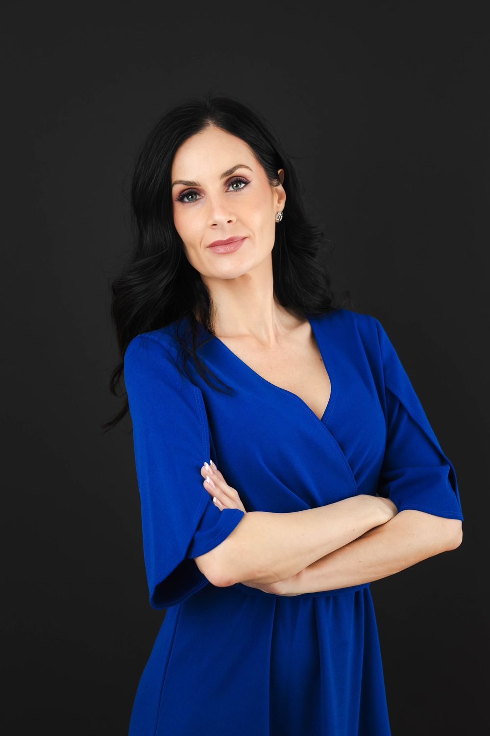 Professional woman in blue wrap dress with arms crossed posing against a dark background.