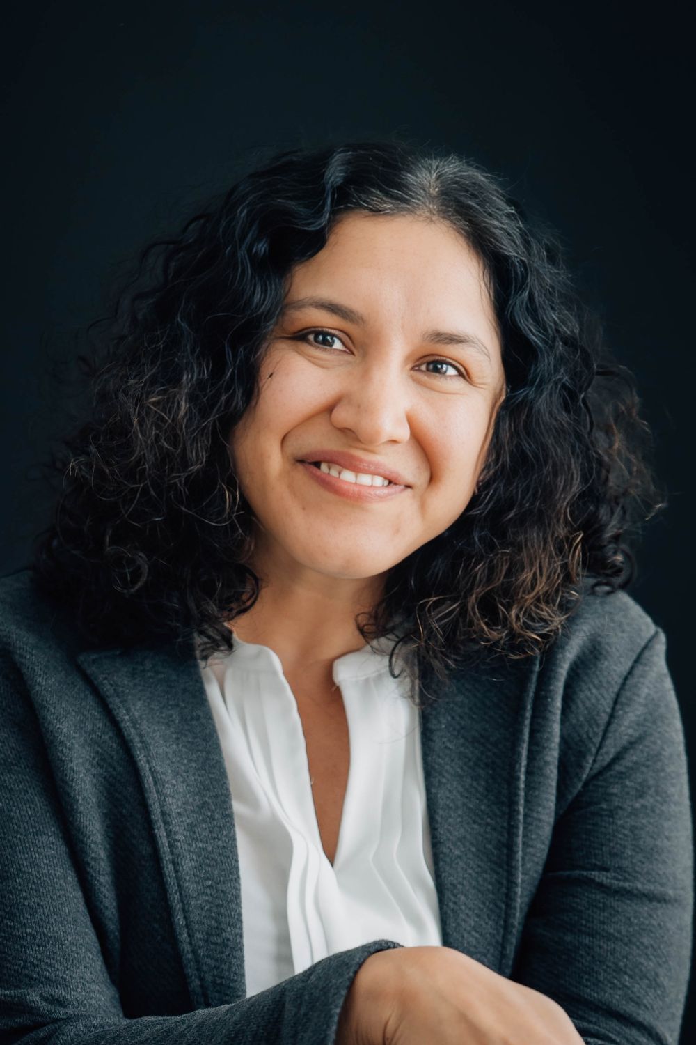 Curly-haired woman in gray blazer smiling with arms crossed against dark background.