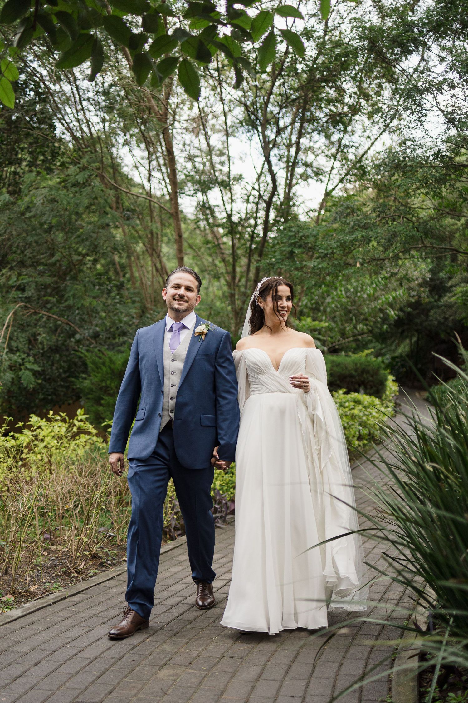 Newlyweds walking together on a wooden boardwalk surrounded by lush garden greenery.