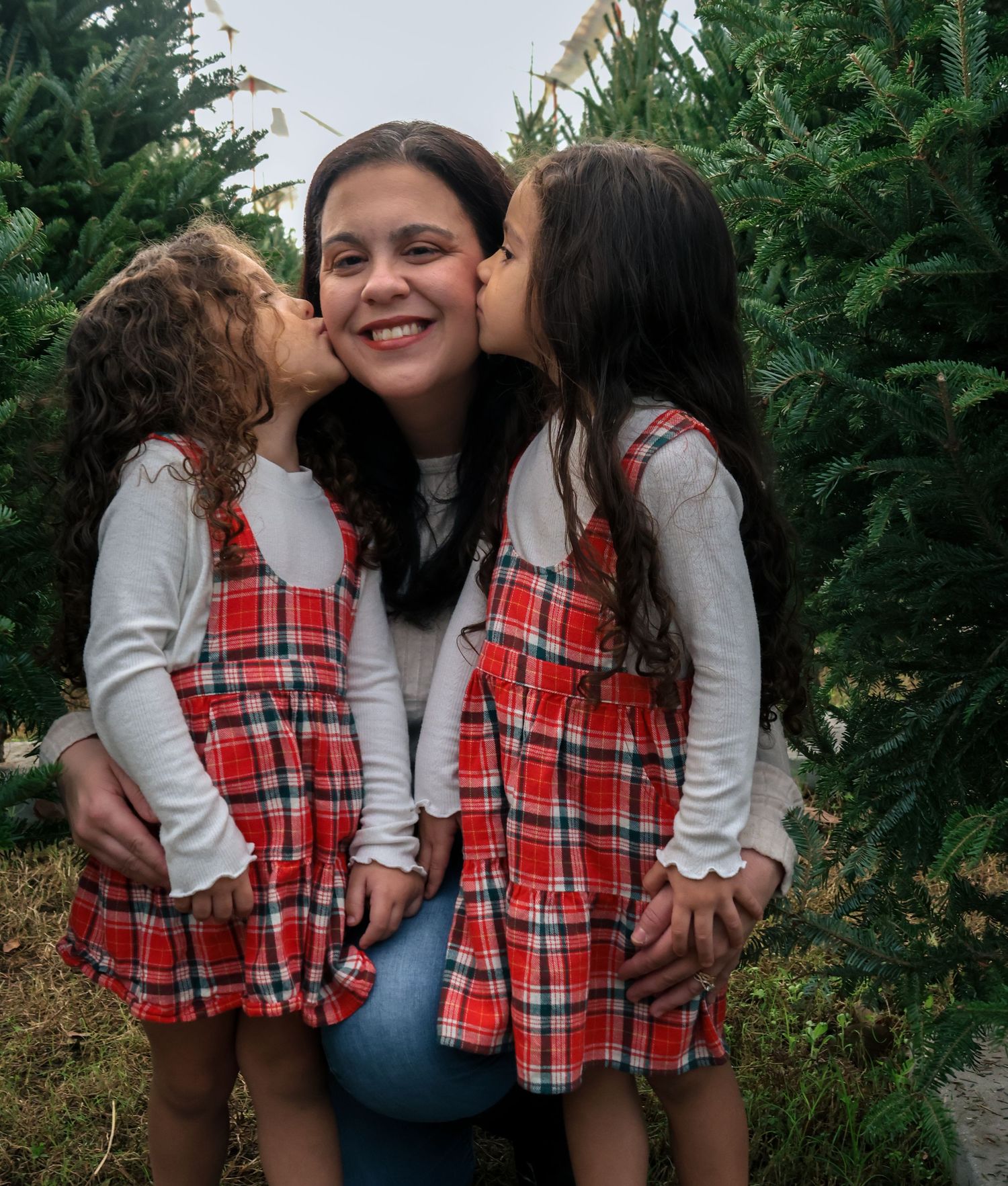 Happy mother smiling as two daughters in matching red plaid dresses kiss her cheeks near Christmas trees.
