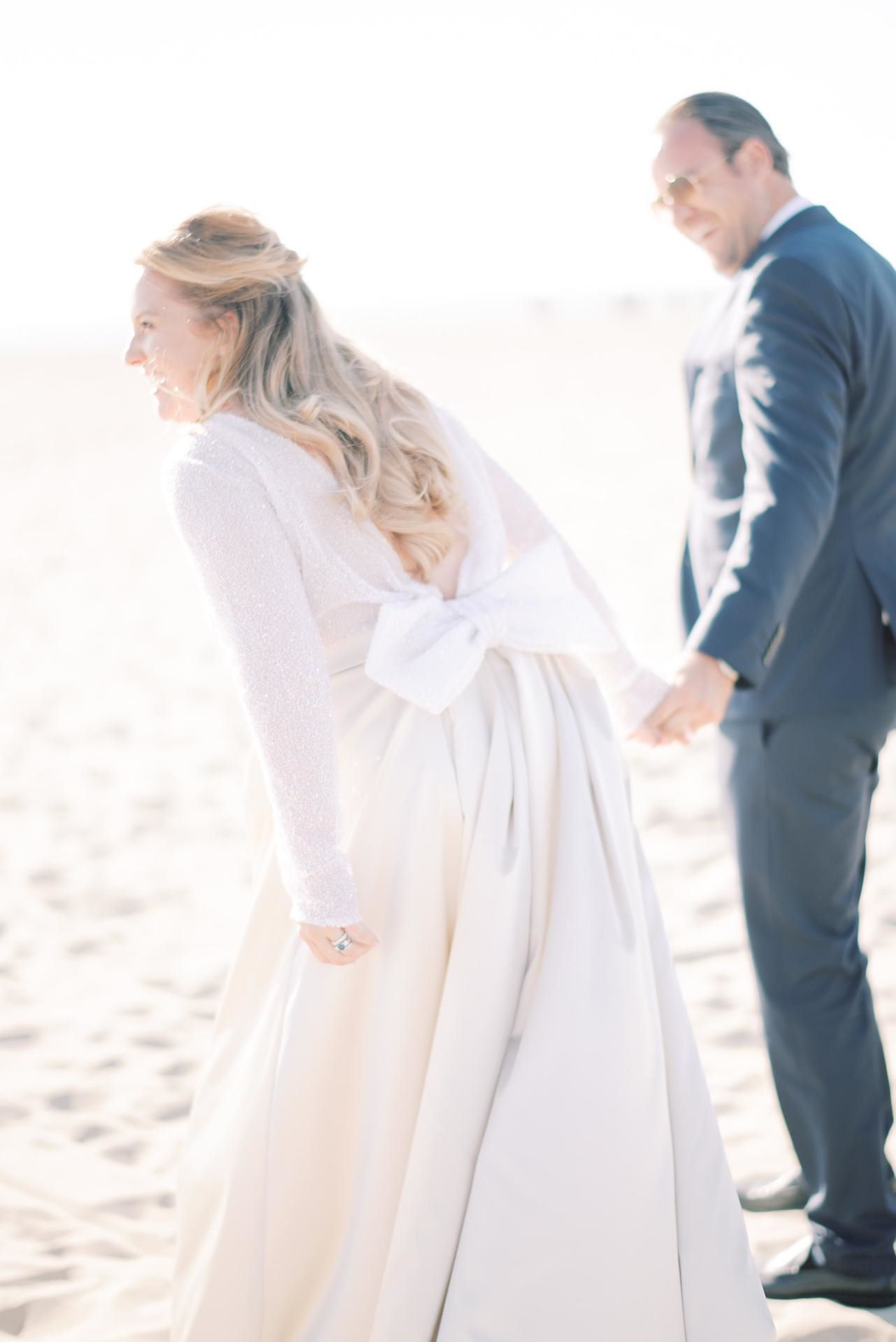 Ein Brautpaar am Strand. Die Braut trägt ein langes Kleid mit einer Masche auf dem Rücken. Der Bräutigam trägt einen blaugrauen Anzug.