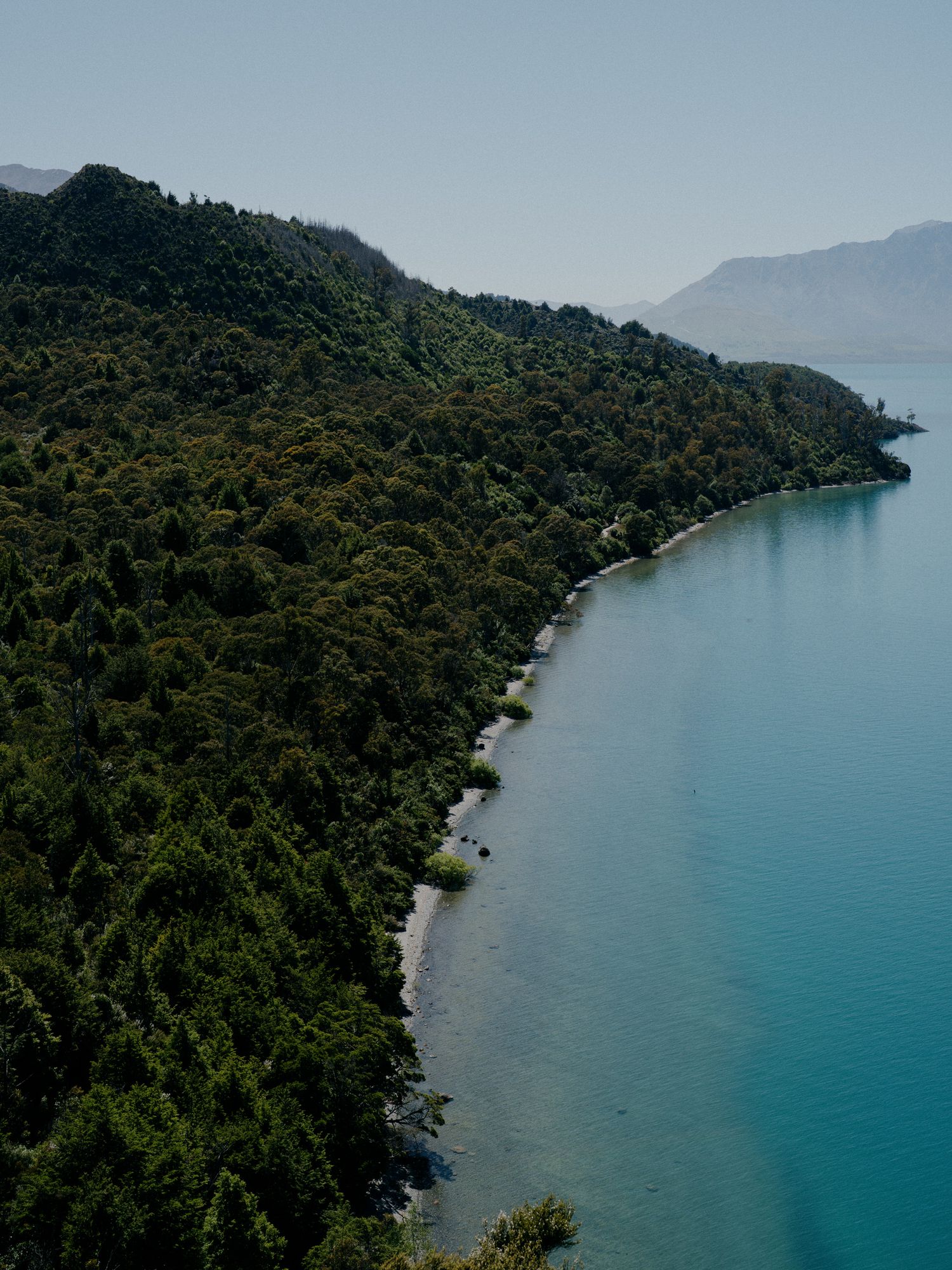 Aerial view of a turquoise coastline with dense forest covering the mountainous shoreline.