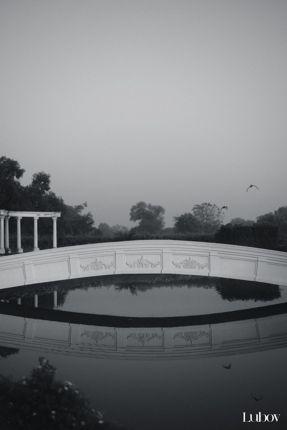 Black and white architectural photograph of curved bridge with classical details reflecting in still water.