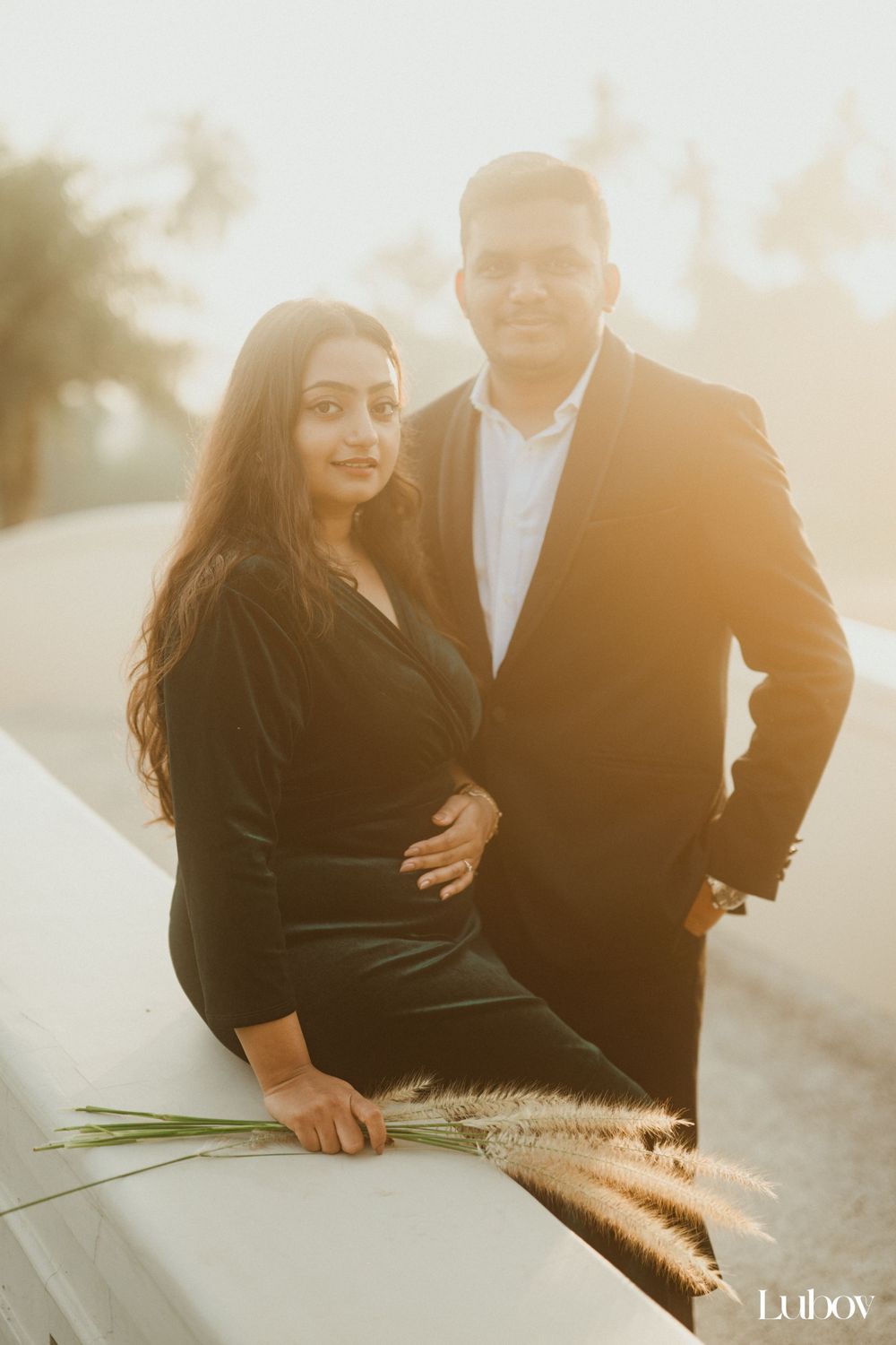 A couple in formal wear poses together on a white surface with decorative pampas grass.