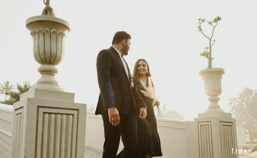Elegant couple walks between decorative stone pillars in warm sunset light.