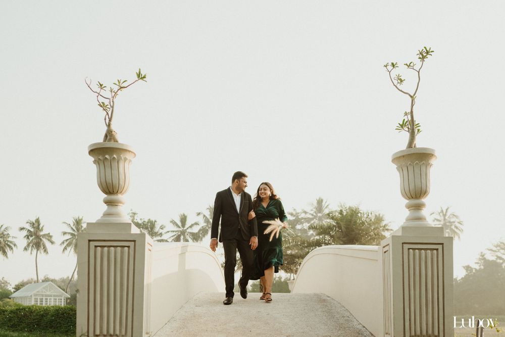 Couple walks between ornate stone urns on a white pathway with palm trees in background.