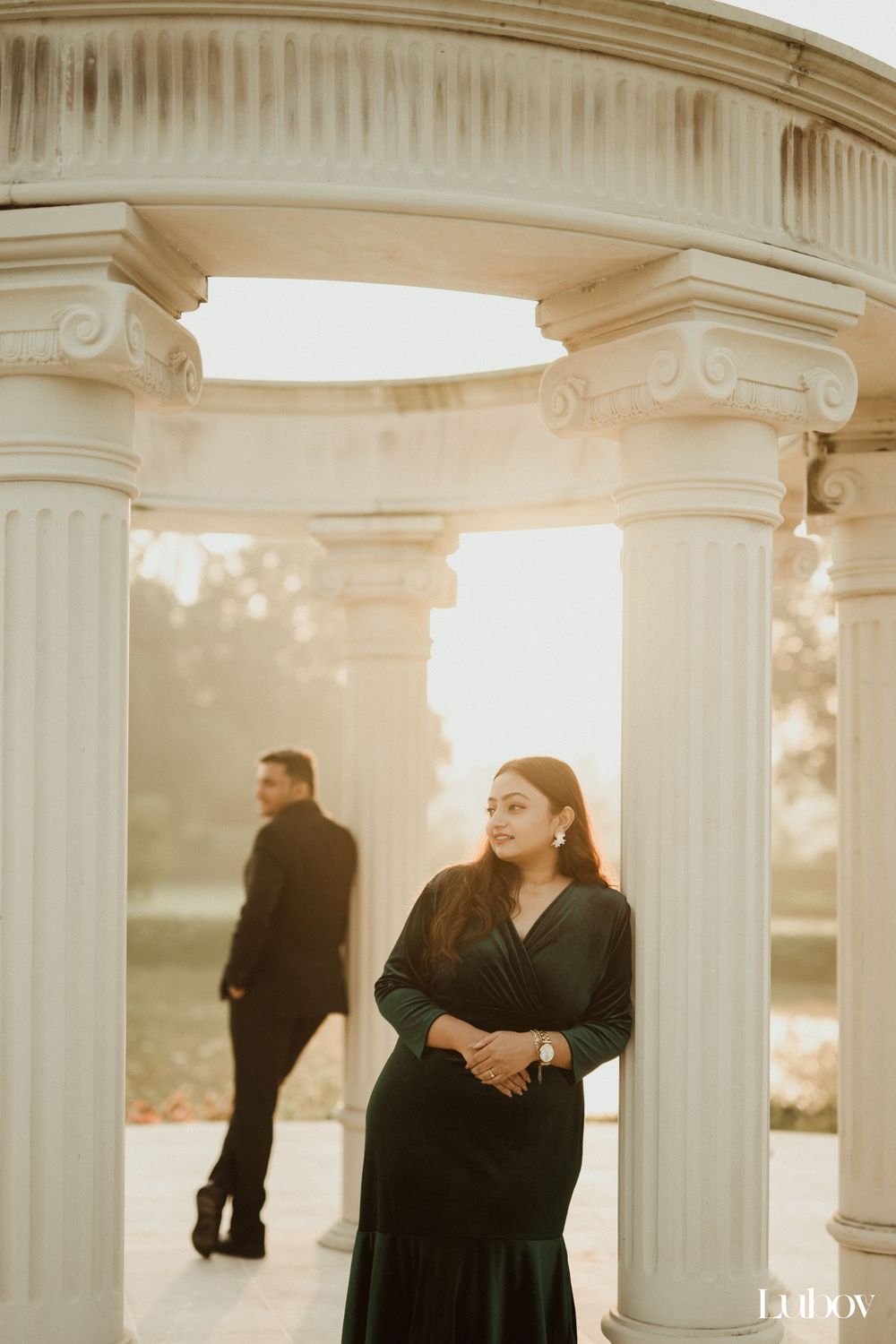 Elegant couple posing separately between white classical columns at sunset.