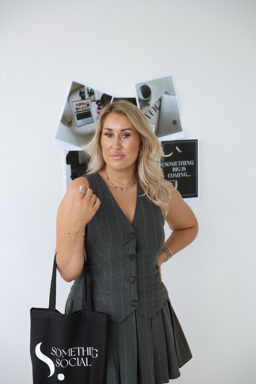 Woman in gray vest dress holding a black tote bag poses against wall with black and white photos.
