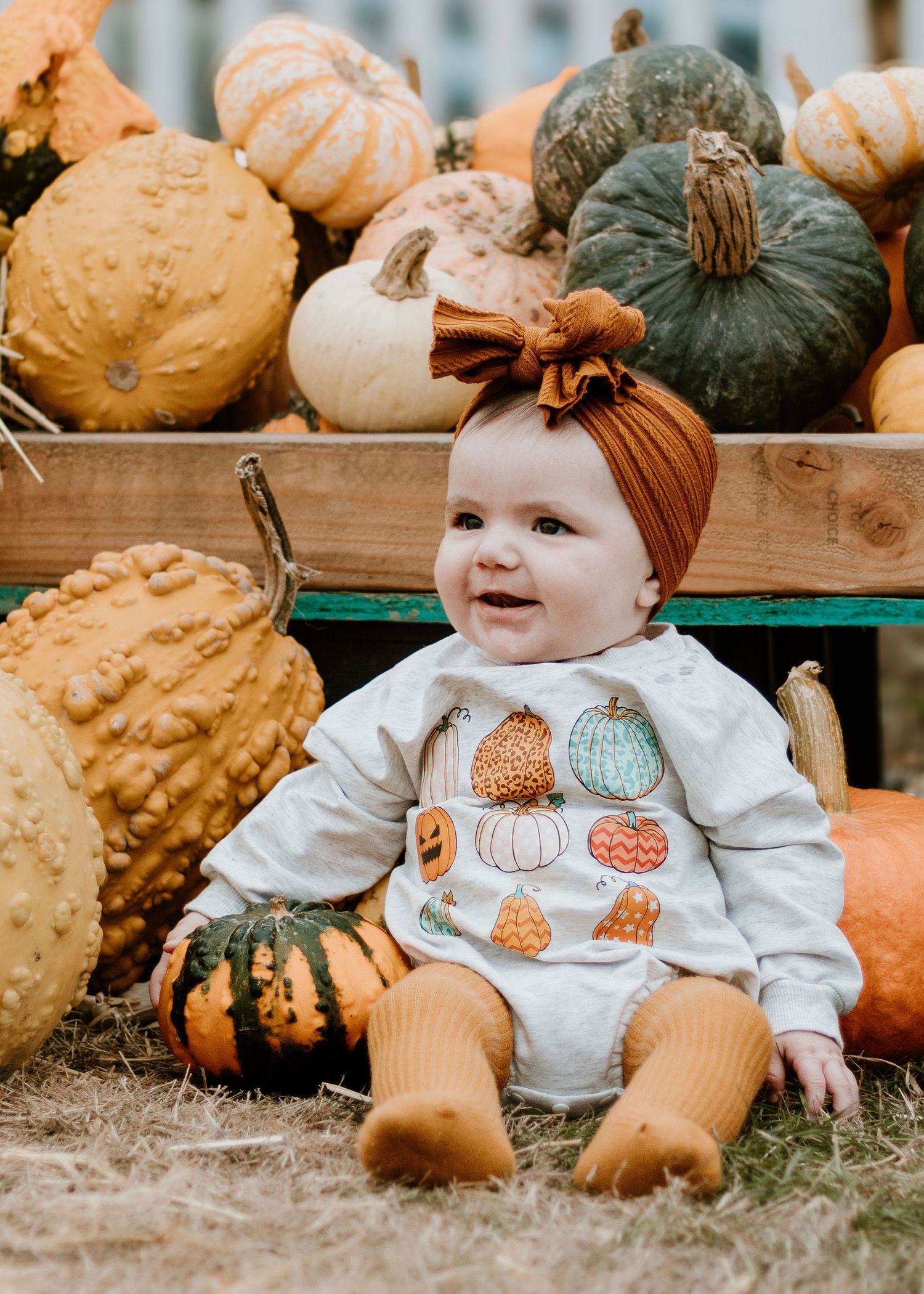 Baby in pumpkin-themed outfit sitting among decorative gourds and pumpkins.
